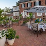 A large red brick house and its back yard. There are several potted plants and trees sitting on the brick patio along with a set table.