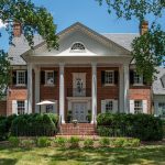 An exterior shot of a luxurious red brick home with white pillars. The image shows off the landscaping around the home.