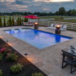 A picture taken at dusk of an inground swimming pool, stone patio, white fence, and countryside. There are various plants and bushes planted around the pool. A table is set with white glasses, napkins, plates, and placemats.