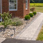 A stone grey pathway in front of a home leading to the back yard. The house is red brick. And there is river rock in the flower beds.