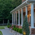 A front porch of a red brick home with white pillars. There is a grey stone pathway leading from the driveway to the front porch. There are flower beds with river rock in front of the porch.