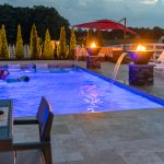 An outdoor living space complete with inground swimming pool, fountains, stone pathways, and luxurious seating. It is dusk outside.