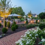 A beautiful backyard with lush green trees, bushes, and white flowers throughout the brick walkways and patio. The trees are illuminated with outdoor lighting fixtures.